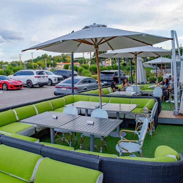 Outdoor restaurant patio with green cushioned seating, tables, umbrellas, and parked cars nearby.