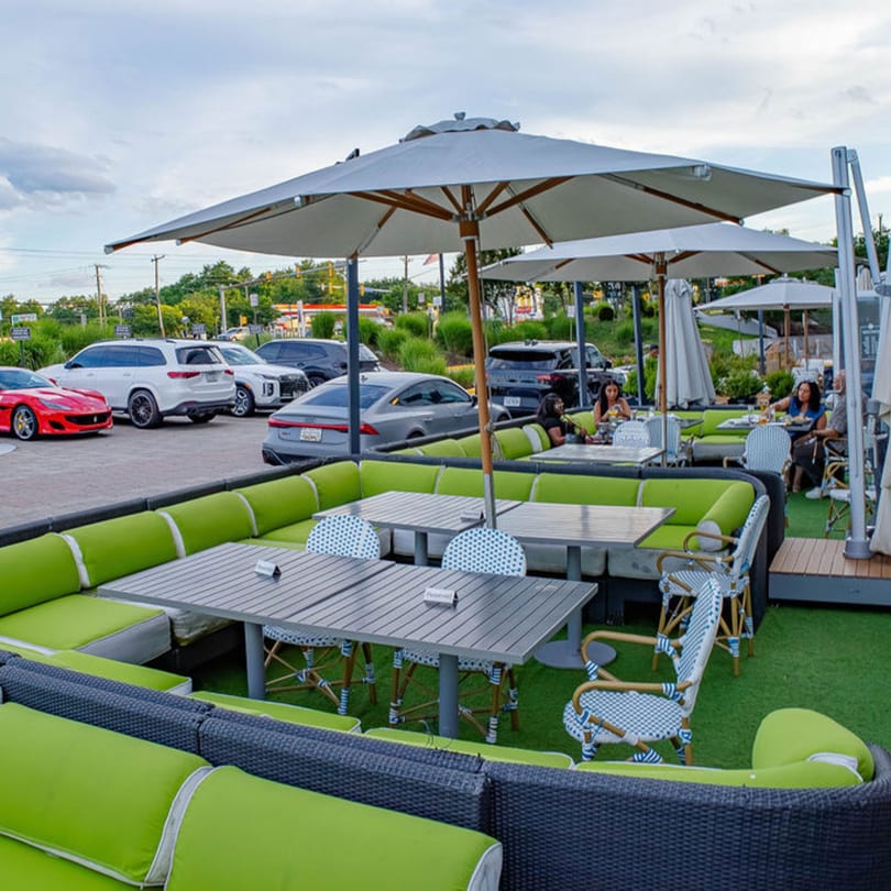 Outdoor restaurant patio with green cushioned seating, tables, umbrellas, and parked cars nearby.