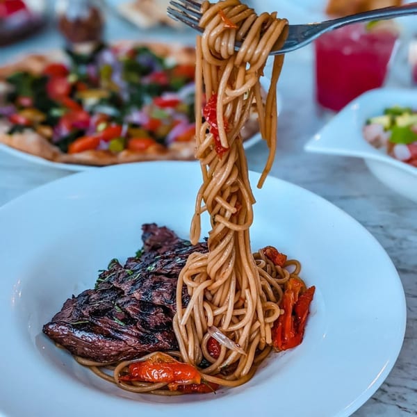 Fork lifting Asian-fusion noodles with red peppers from a plate with steak and other dishes.