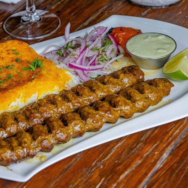 Two skewers of grilled meat, rice, red onions, roasted tomatoes, and green sauce on a white plate.