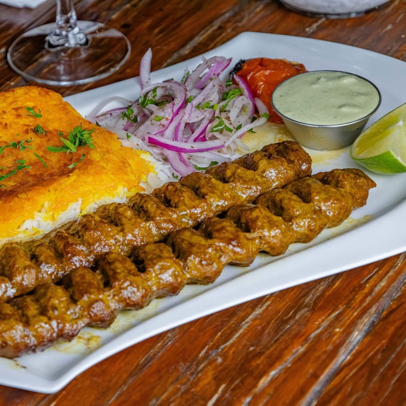 Two skewers of grilled meat, rice, red onions, roasted tomatoes, and green sauce on a white plate.