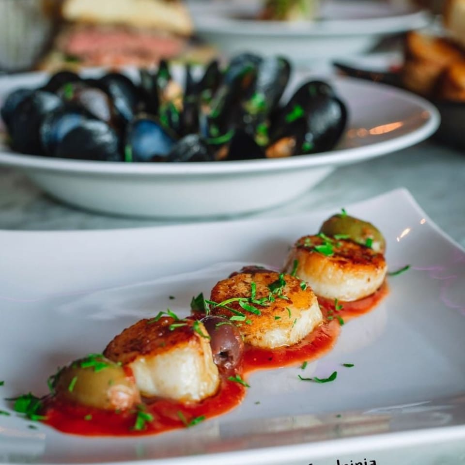 Seared scallops with red sauce and olives, garnished with herbs, with mussels in the background.