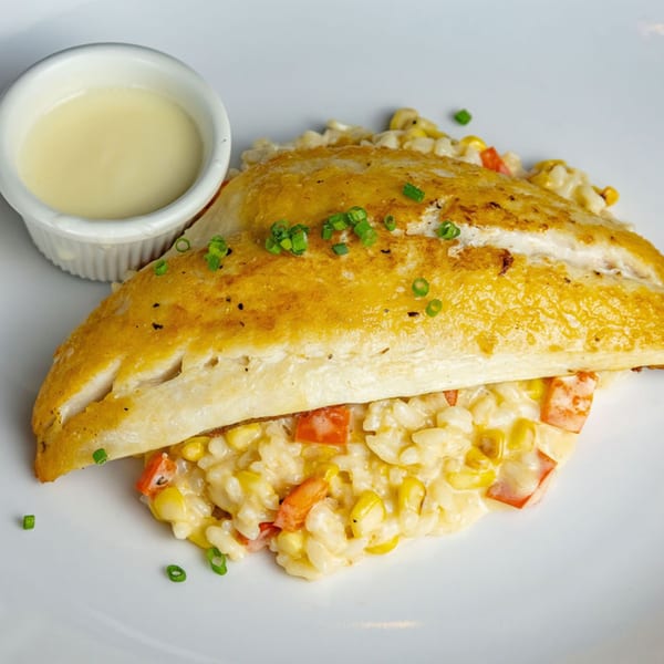 Golden-brown fish fillet over creamy risotto with corn and red peppers, served with a side of sauce.
