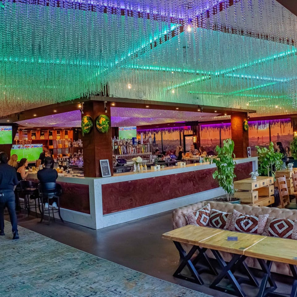 A vibrant bar and lounge with a glowing green and purple ceiling, patrons at the bar, and lounge seating.