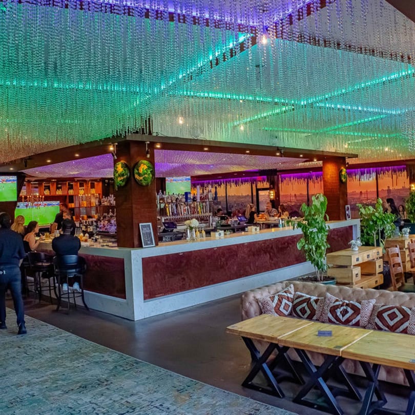 A vibrant bar and lounge with a glowing green and purple ceiling, patrons at the bar, and lounge seating.
