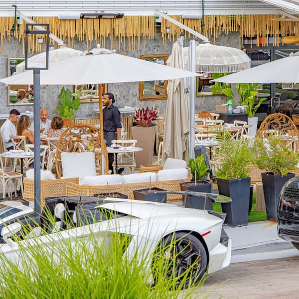 Outdoor restaurant patio with diners, white umbrellas, lush plants, and a white sports car parked nearby.