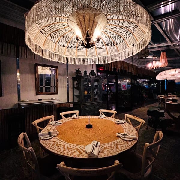 A round dining table with place settings under a large, decorative woven chandelier in Greenhouse Kitchen + Bar.
