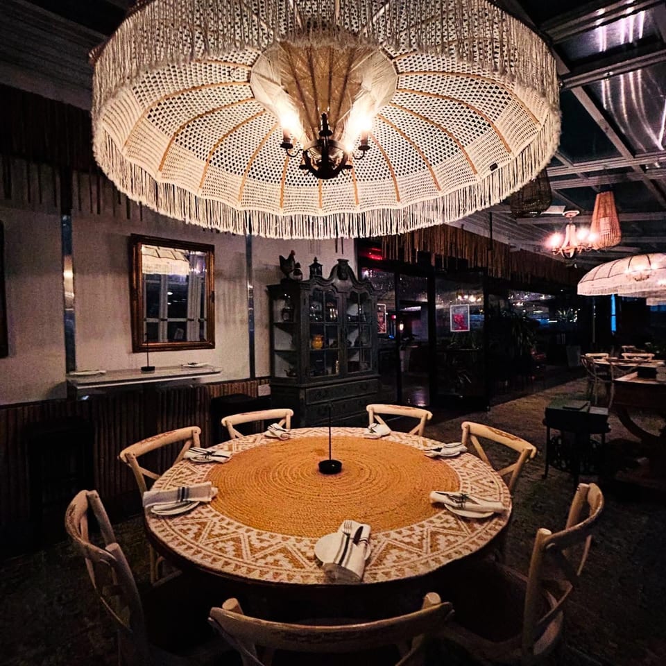 A round dining table with place settings under a large, decorative woven chandelier in Greenhouse Kitchen + Bar.