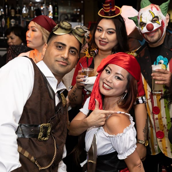 Four people in Halloween costumes, including a pirate, ringmaster, and clown, pose with drinks at Greenhouse Kitchen + Bar.