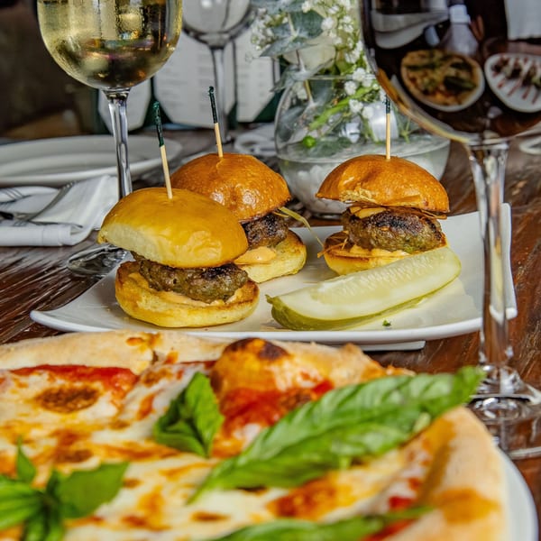 Three mini burgers with a pickle spear and a pizza slice on a wooden table.