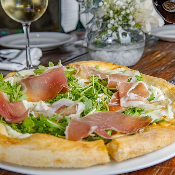 A white pizza topped with prosciutto, arugula, and ricotta cheese, with a glass of white wine.