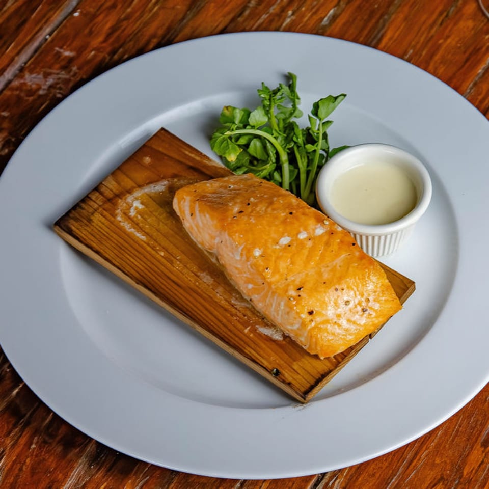 Grilled salmon fillet on a wooden plank, served with fresh greens and a side of sauce.