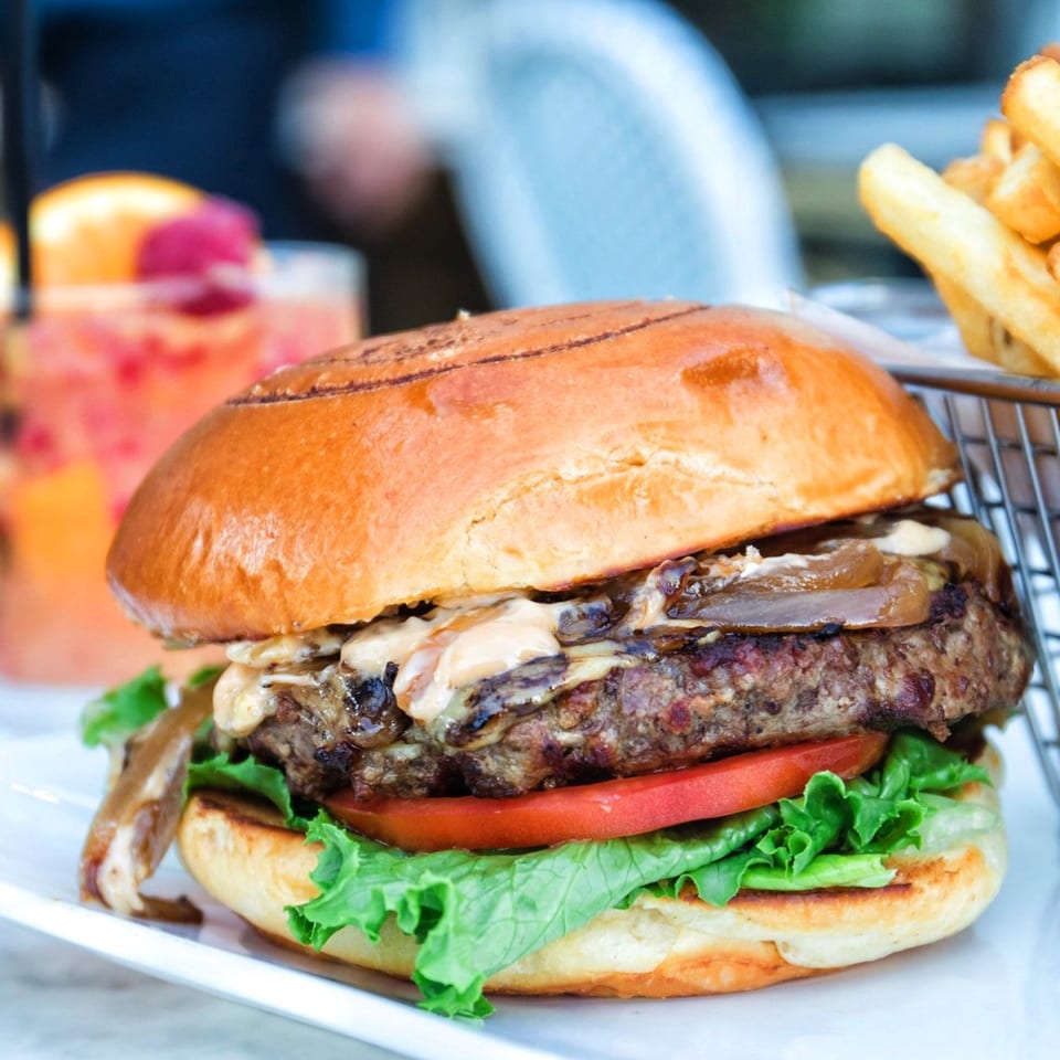 Gourmet burger with caramelized onions, cheese, lettuce, and tomato, served with fries and a cocktail.